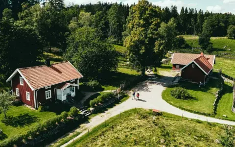 Två röda hus med tegeltak vid Rya Åsar, Borås. En grusväg leder förbi husen genom ett grönskande landskap med skog och ängar.
