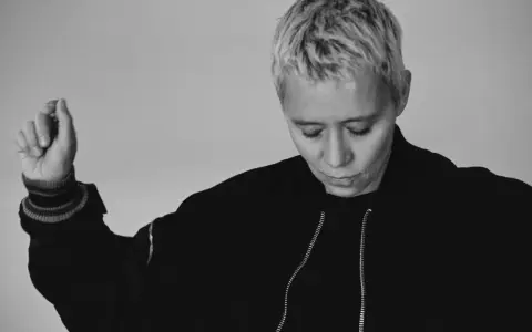 Jennifer Cardini, con i suoi capelli biondi corti e un'espressione pensierosa, alza un braccio con un gesto delicato. Indossa una giacca scura con zip.
