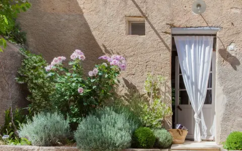 Lavendel beet ideen: Ein mediterraner Garten mit Rosen, Lavendel und einer Tür mit weißem Vorhang.