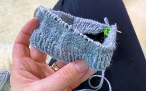En hand håller ett stickat provstycke i ljusgrått garn. Tekniken german short rows används för att forma det.