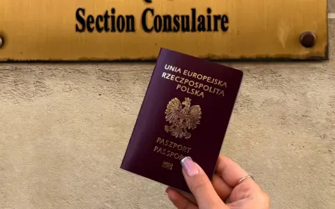 Dłoń trzyma polski paszport na tle tablicy z napisem "Section Consulaire".
