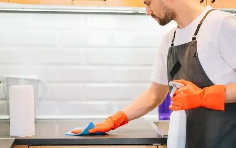 Hombre con guantes naranjas desinfecta la cocina, rociando la encimera con un spray limpiador.