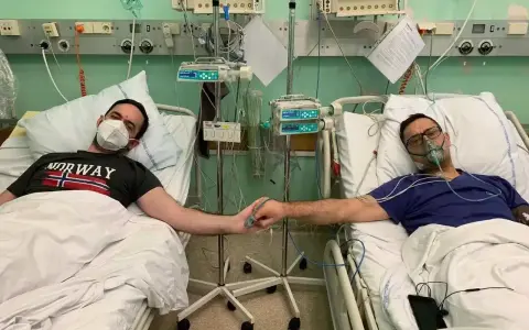 Dwóch mężczyzn w szpitalnych łóżkach, po operacji guza nerki, trzyma się za ręce. Jeden ma maskę tlenową, drugi zakrywa usta.
