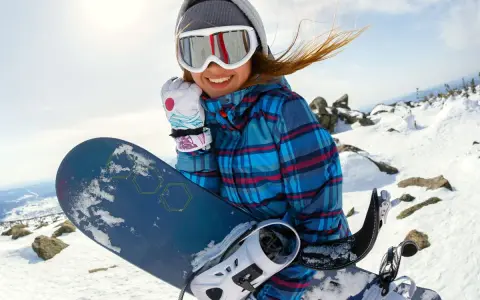 Uśmiechnięta snowboardzistka w goglach i czapce trzyma deskę snowboardową na tle zaśnieżonych gór.
