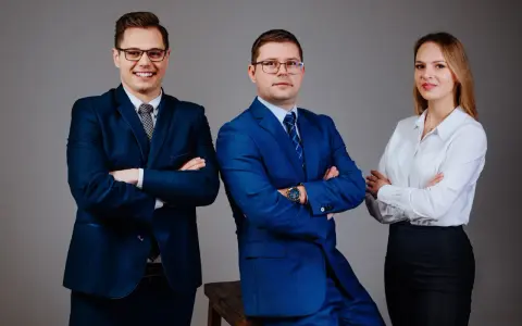 Trzy osoby w garniturach i białej koszuli, stojące na szarym tle, z rękami skrzyżowanymi na piersi.