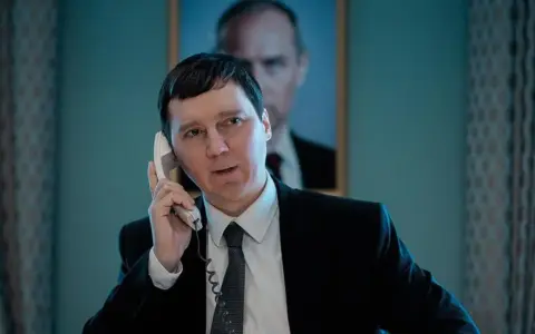 Un uomo in giacca e cravatta parla al telefono, con un ritratto di Putin sullo sfondo. Sembra il mago del Cremlino.