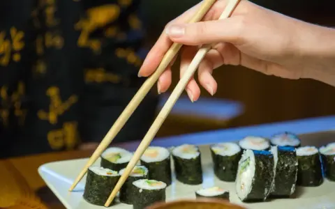Dłoń z pałeczkami sięga po rolkę sushi. Na talerzu ułożone są kawałki sushi, każdy z nich to mała porcja smaku i kalorii.