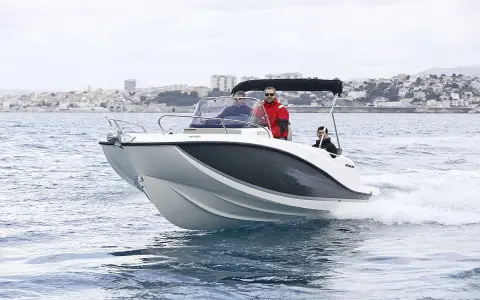 Bateau blanc et gris naviguant sur l'eau, avec trois personnes à bord. Le tableau de bord indique la puissance du moteur du bateau. Ville en arrière-plan.