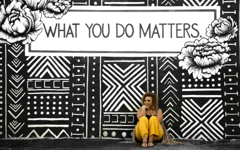 Kobieta siedzi przed muralem z napisem "WHAT YOU DO MATTERS". To inspirujący dialog motywujący.