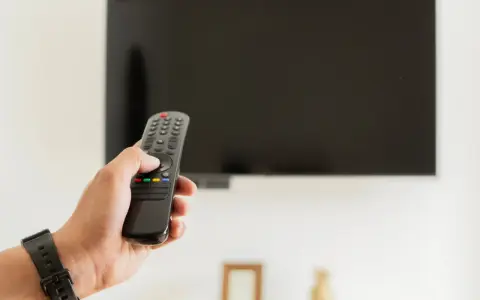 Dłoń trzymająca pilot do telewizora na tle wyłączonego ekranu.