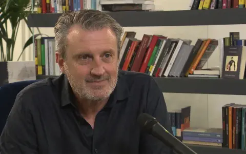 Uomo con barba grigia sorride, microfono davanti. Alle sue spalle scaffali pieni di libri, tra cui "Le ossa parlano trama".