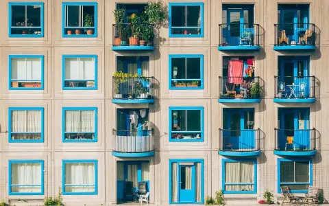 Fachada de edificio con ventanas y balcones de marcos azules. Cada balcón muestra detalles de la vida cotidiana, como plantas y ropa tendida, reflejando los gastos de comunidad.