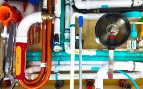 Un réseau complexe de tuyaux blancs, orange et turquoise, avec un outil de plomberie et un manomètre. Un **profilé souple** orange se courbe gracieusement.