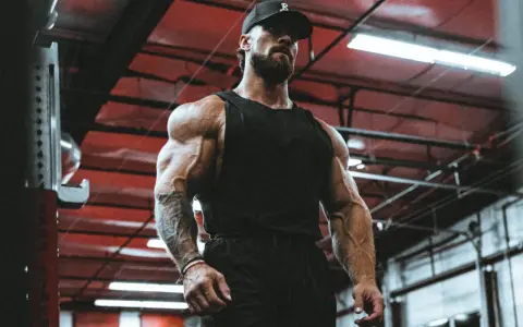 Ein Mann mit beeindruckendem **mens physique** steht im Fitnessstudio. Seine Muskeln sind stark definiert, er trägt eine schwarze Kappe und ein Tanktop.