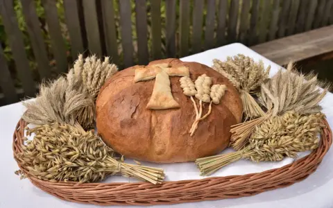 Chleb dożynkowy ozdobiony krzyżem i kłosami, otoczony snopkami zbóż. Inspiracja, jak ozdobić chleb dożynkowy.