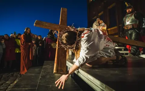 Scena drogi krzyżowej: Jezus w cierniowej koronie, z krwią na szacie, upada pod ciężarem krzyża. Tłum widzów obserwuje to poruszające przedstawienie.