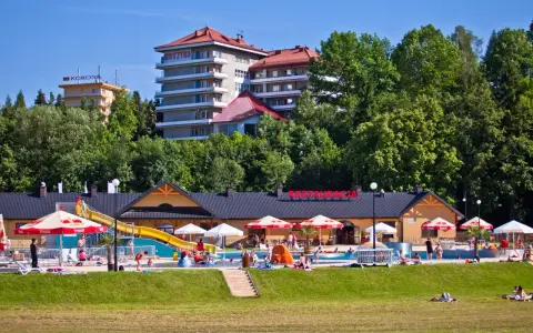 Słoneczny dzień w Muszynie-Zdroju. Ludzie relaksują się przy basenie zjeżdżalnią, a w tle widać hotel i restaurację.