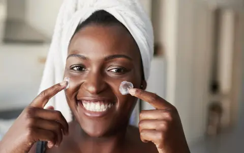 Sourire radieux, une femme applique une crème sur ses joues. Un avant-après prometteur pour une peau éclatante grâce au rétinol.