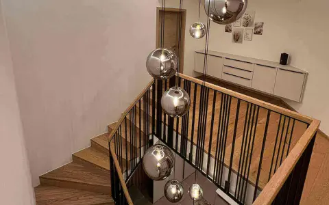 Nowoczesna klatka schodowa w domu z drewnianymi stopniami i czarną balustradą, ozdobiona wiszącymi, lustrzanymi kulami.