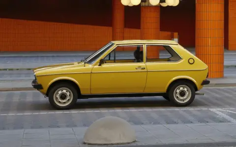 Żółty Audi 50, klasyk z lat 70., parkuje na tle pomarańczowej, kafelkowej architektury.