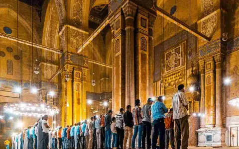 Männer beten Tarawih in einer Moschee. Sie stehen in Reihen vor kunstvoll verzierten Säulen und Bögen, beleuchtet von zahlreichen Lampen.
