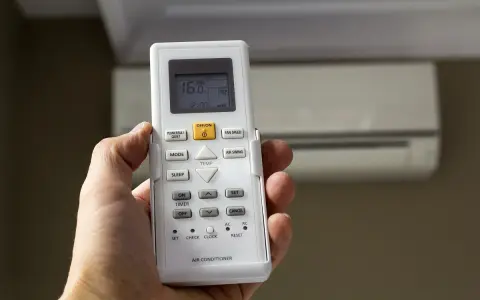 Mano sujetando mando para configurar mando universal aire acondicionado sin código. Aire acondicionado al fondo.