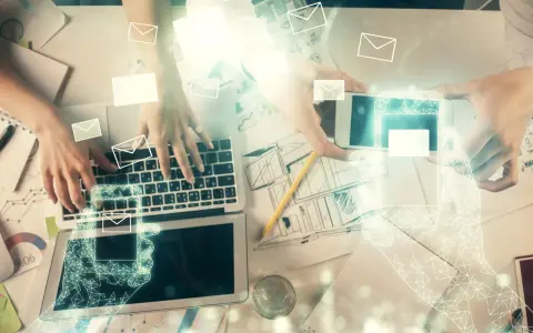 Dwie osoby pracują przy biurku, jedna pisze na laptopie, druga trzyma smartfon. Wokół latają ikony kopert, sugerując komunikację. Jak zacząć maila?