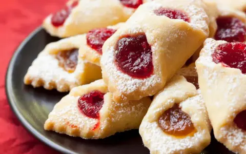 Ciastka kruche con ripieno di marmellata, spolverati di zucchero a velo, su un piatto scuro.