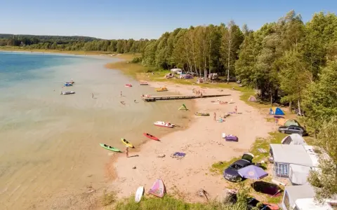 Letnie popołudnie nad jeziorem Świętajno. Ludzie kąpią się, pływają kajakami i odpoczywają na plaży wśród namiotów i kamperów.