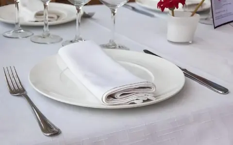 Une table dressée avec une assiette blanche, une serviette de table pliée, une fourchette et un couteau. La dimension serviette de table est parfaite pour cette présentation élégante.