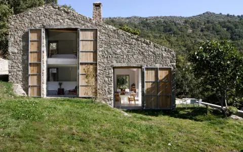 Una casa in pietra con grandi porte in legno, perfetta per chi ama vivere in campagna. Si intravedono interni moderni e un paesaggio collinare rigoglioso.