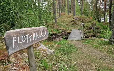 Träskylt med texten "Flottarleden Munkedal" pekar mot en liten träbro över en bäck i en skogsmiljö.