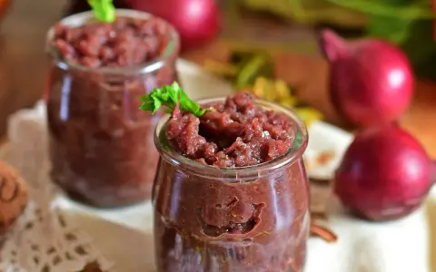 Dwa słoiczki z domowym, aromatycznym chutney z czerwonej cebuli, ozdobione listkiem zieleniny.
