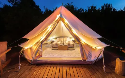 Mysig glamping i Sverige. Ett upplyst tält med säng och mysig inredning på en träaltan, omgivet av mörka träd.