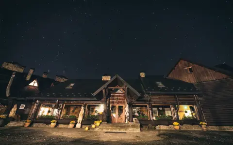 Nocne niebo nad bacówką Biały Jeleń. Drewniany budynek z oknami oświetlonymi ciepłym światłem, otoczony donicami z kwiatami.