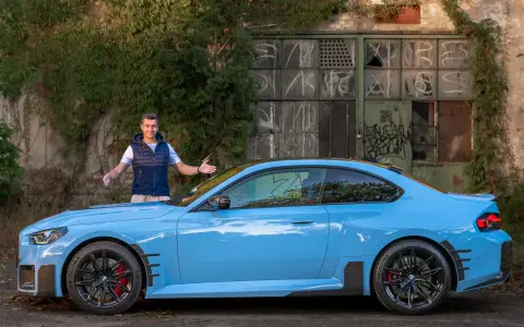 Mężczyzna prezentuje błękitne BMW M2 G87 przed zniszczonym budynkiem.