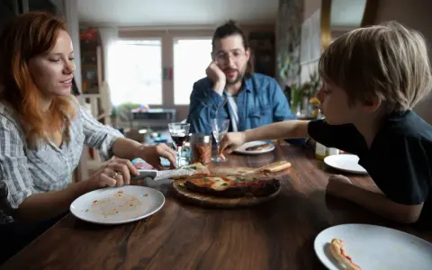 Rodzina przy stole, pizza, w tle mężczyzna zamyślony. Czasem nawet przy wspólnym posiłku pojawiają się kłótnie o dzieci z poprzedniego związku.