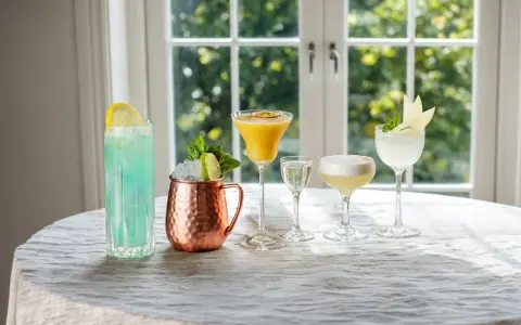 Fem olika vodka drinkar, från en blå long drink till en gyllene passionfruktscocktail och en vit äppeldryck, står uppradade på ett dukat bord.
