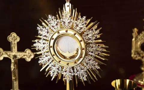 A golden monstrance holds the blessed sacrament, surrounded by ornate details and red jewels. A crucifix stands nearby, a symbol for a blessed sacrament prayer request.