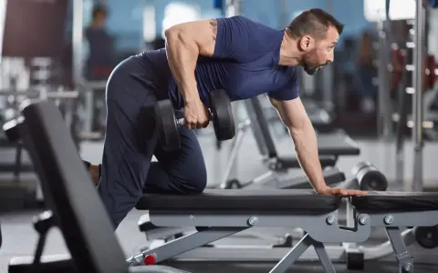 Man utför enarms hantelrodd på ett gym. Han stöttar sig mot en bänk med ena handen och drar en hantel mot bröstet med den andra.