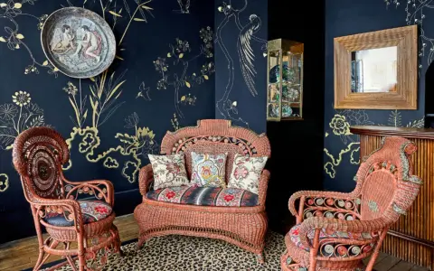 Salon au style napoléon 3, avec des fauteuils et un canapé en rotin corail, sur un tapis léopard, devant un papier peint bleu nuit orné de motifs dorés.