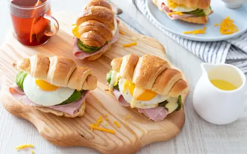 Trois croissants au jambon, œuf au plat, avocat et fromage râpé, servis sur une planche en bois avec une tasse de thé.