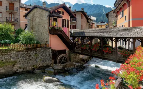 Un pittoresco ponte di legno attraversa un torrente impetuoso, fiancheggiato da case colorate e fiori rossi. Cosa vedere in questo incantevole borgo alpino.