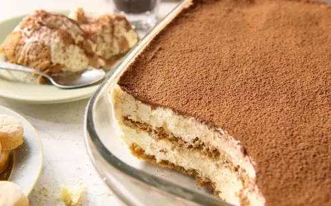 Pyszne tiramisu bez jajek, obficie posypane kakao, w szklanej formie. Obok porcja na talerzyku.