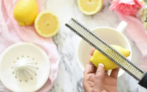 Hand reibt zeste einer zitrone über eine Reibe. Zitronen und Blumen liegen auf einem Marmortisch.