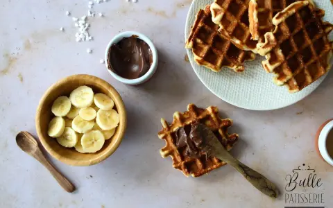 Une gaufre bubble est garnie de pâte à tartiner au chocolat. À côté, des rondelles de banane et une cuillère en bois.