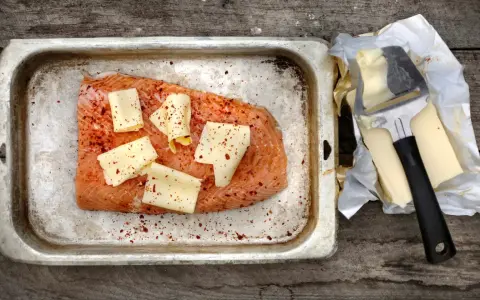 Lax i ugn, kryddad med chili och toppad med ostskivor, redo att tillagas. Bredvid ligger smör och en osthyvel.