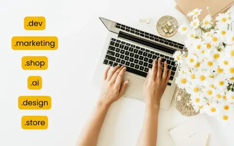 Kobieta pisze na laptopie, obok bukiet margaretek. Widoczne są różne rozszerzenia domen, np. .dev, .marketing, .shop, .ai, .design, .store.