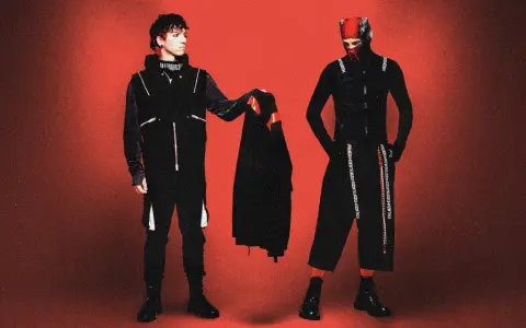 Dwóch artystów w czarnych strojach, przypominających styl z okładek płyt twenty one pilots, pozuje na tle czerwonym.