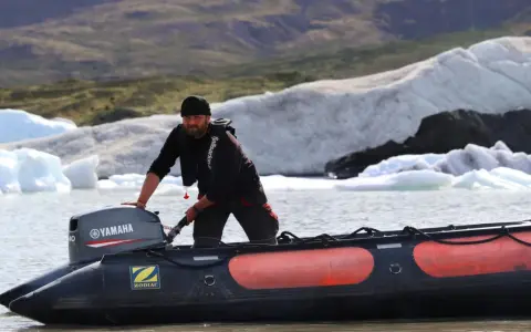 Un homme tente de démarrer un moteur 2 temps sur un Zodiac, mais il cale. Derrière lui, des icebergs flottent dans un lac glaciaire.